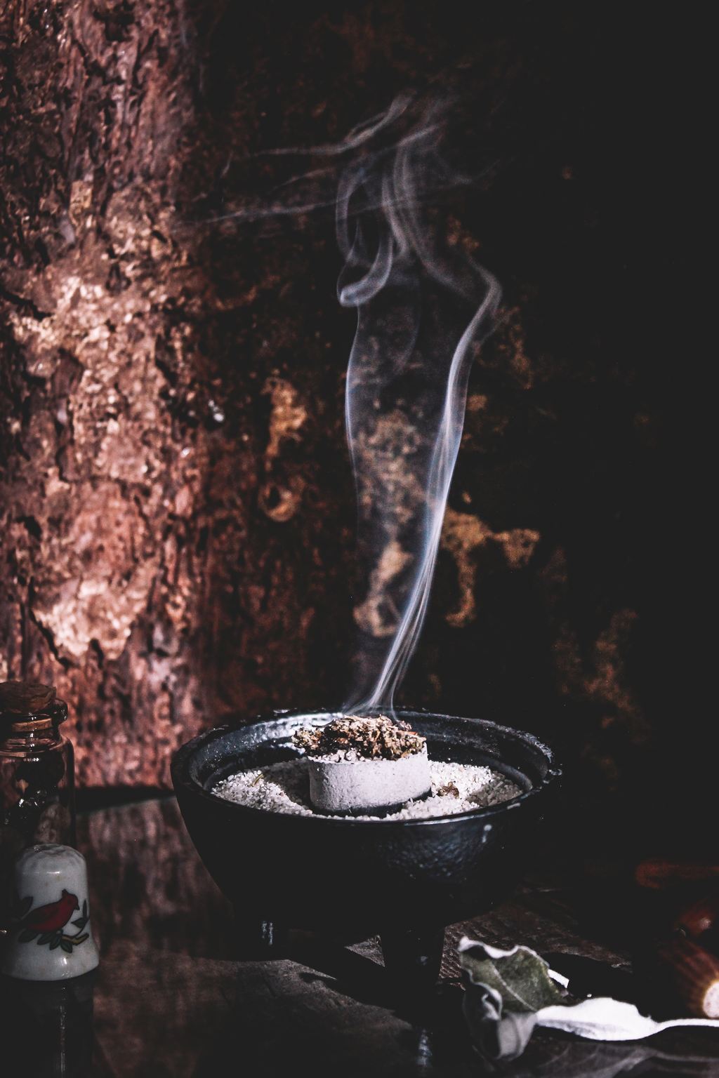 Ein Räuchergefäß aus Gußeisen mit weißem Sand gefüllt. Darauf liegt ein Stück gut durchgeglühte Räucherkohle auf der rauchendes Kraut gelegt ist. Der Rauch steigt nach oben.