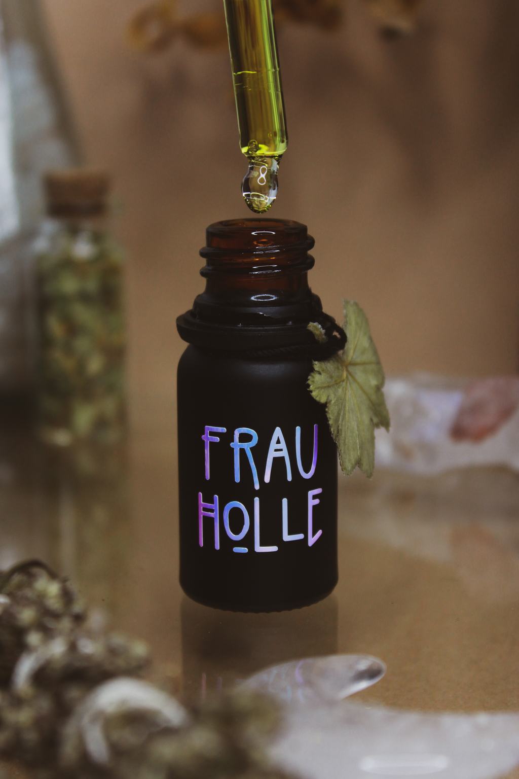 Eine mattschwarze Pipettenflasche mit der Aufschrift "Frau Holle". Als Anhänger ist ein Blatt Frauenmantel an der Flasche zu finden.