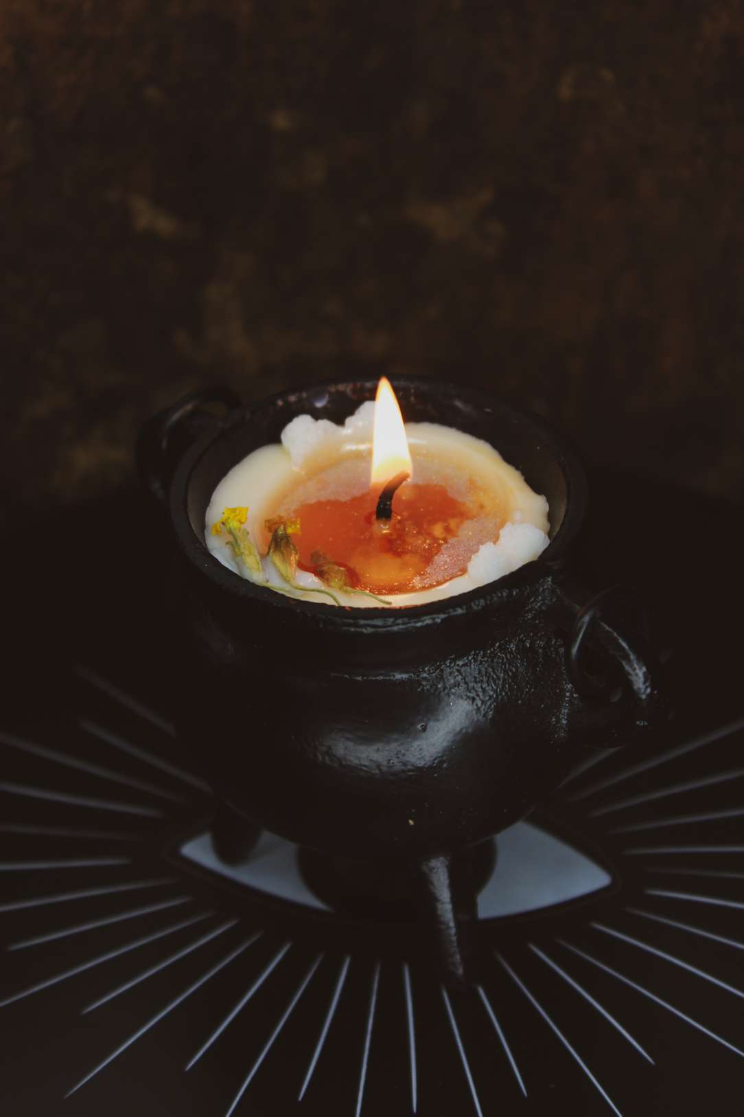 Eine weiße Kerze in einem gußeisernen Hexenkessel. Es sind weiße Schneeflocken aus Wachs darauf drapiert und Schlüsselblumen-Blüten liegen darauf. Die Kerze brennt und unter dem weißen Wachs kommt orangenes zum Vorschein.