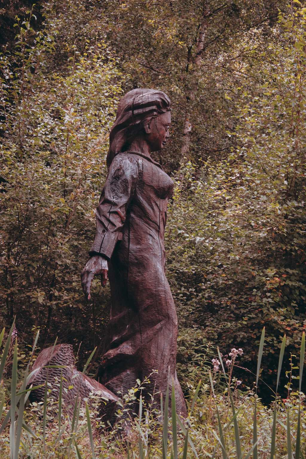 Die Frau-Holle-Figur aus Holz am Frau-Holle-Teich. Umrandet von Gräsern, Kräutern und Bäumen. Ihr Blick ist von uns aus nach rechts gewendet.