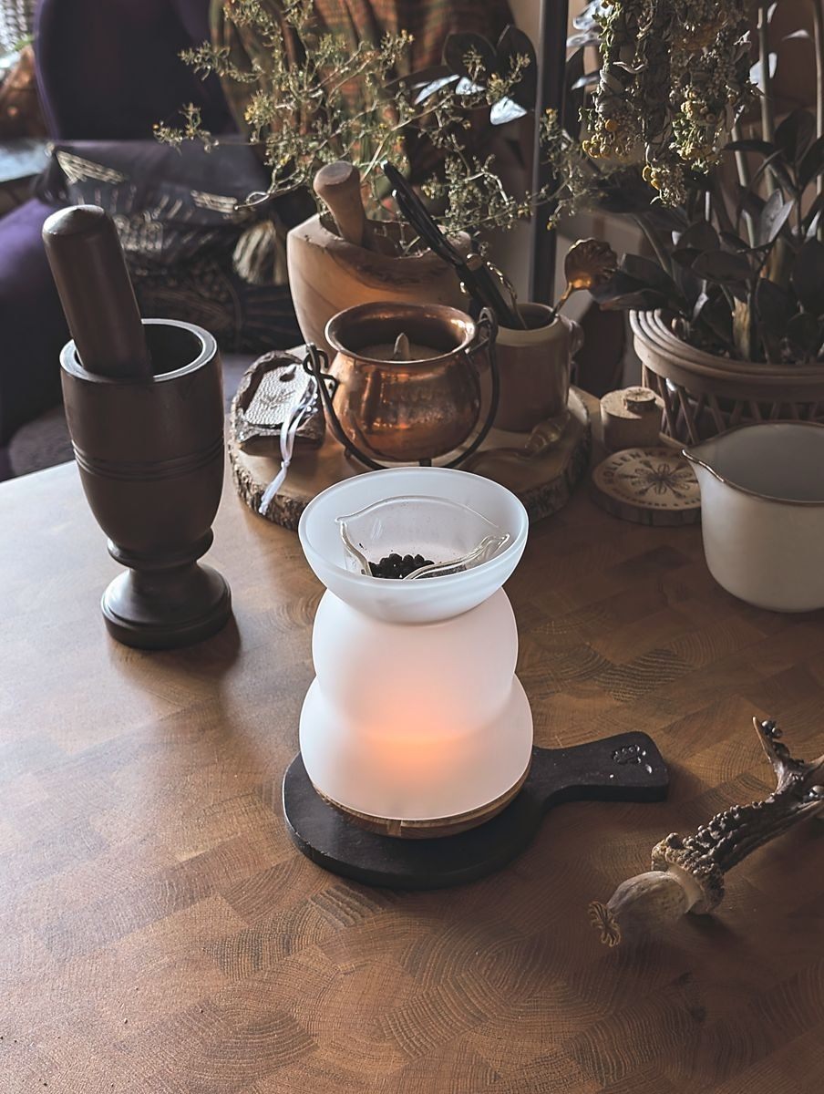 Ein rustikales, gemütliches Arrangement auf einem Holztisch mit einem beleuchteten Räucher-Stövchen aus Milchglas in dem dunkle Beeren liegen, einem dunklen Mörser mit Stößel, einem Kupferkessel und getrockneten Kräutern. Dekorative Elemente wie ein Holztablett, ein kleines Keramikgefäß und ein geschnitztes Geweih ergänzen die Szene. Im Hintergrund sind Pflanzen und ein mit Stoff bezogener Stuhl zu sehen – eine warme, naturverbundene Atmosphäre.