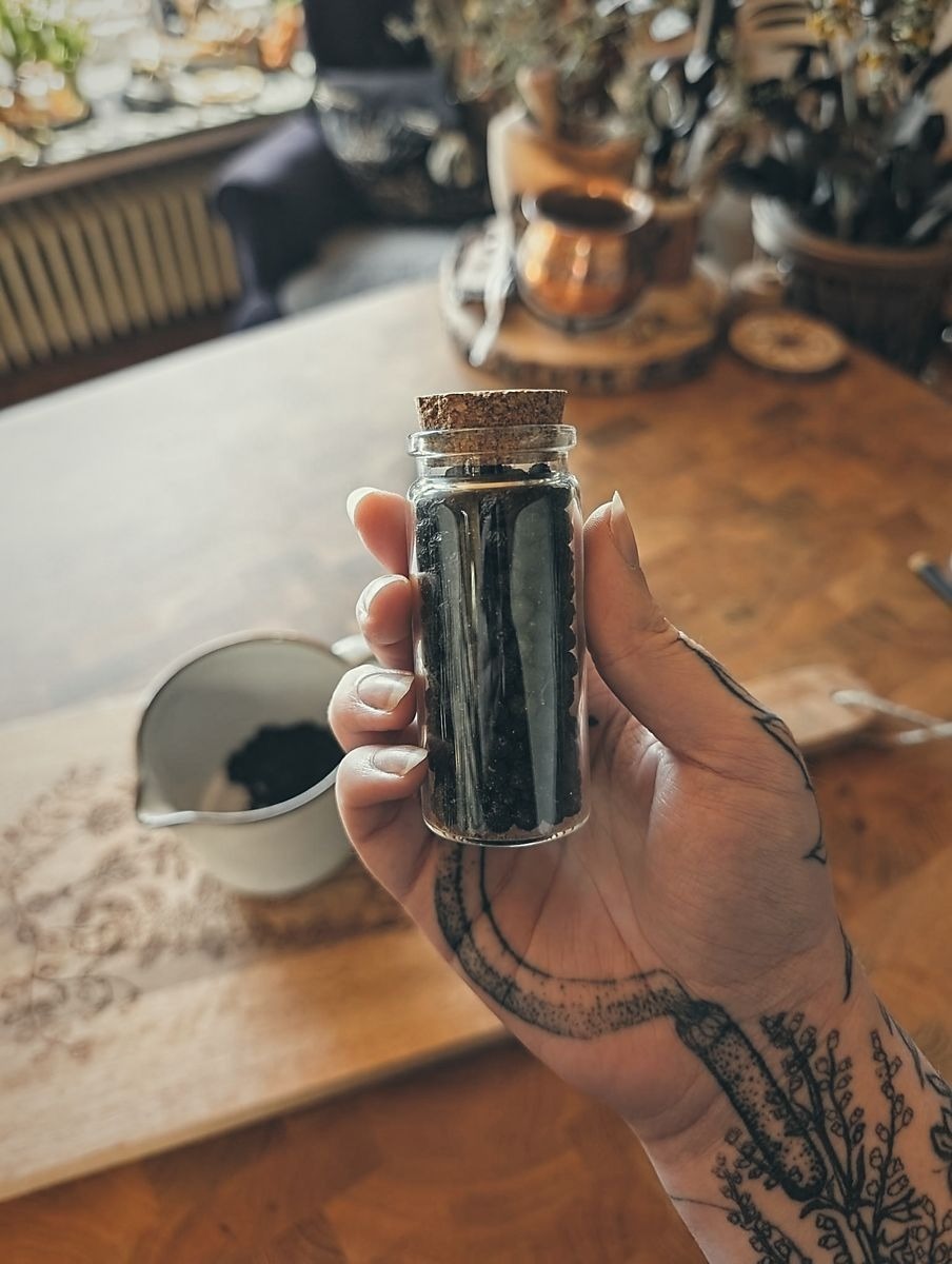 Eine tätowierte Hand hält ein kleines Glasfläschchen mit Korkverschluss, gefüllt mit dunklen Hartriegel-Früchten. Im Hintergrund steht eine weiße Schale mit gleichem Inhalt auf einem Holztisch, umgeben von Kupfergefäßen, Pflanzen und weiteren rustikalen Gegenständen. Die Szene wirkt wie ein liebevoll eingerichteter, naturverbundener Arbeitsbereich mit warmem, handwerklichem Flair.