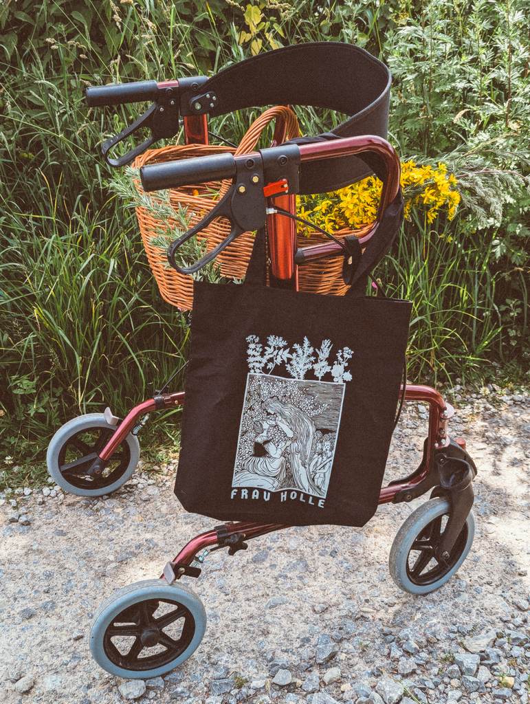 Ein roter Rollator auf dem ein Korb mit Kräutern steht. An einem der Griffe hängt eine Tasche mit Frau Holle Motiv.