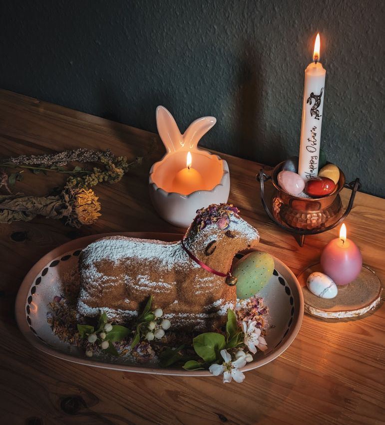 Ein Kuchen in Form eines Lamms, dekoriert mit verschiedenen Blüten und einem roten Band mit einem Glöckchen. Drumherum stehen drei brennende Kerzen und Dekoration wie Eier aus Steinen.