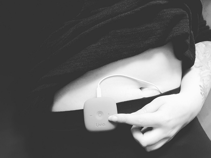 Ein schwarz-weiß Foto das ein TENS-Gerät mit der Aufschrift "Livia" zeigt, das an eine Hose geklippt ist. Eine Hand in an das Gerät gelegt und drückt mit einem Finger die Plus-Taste.