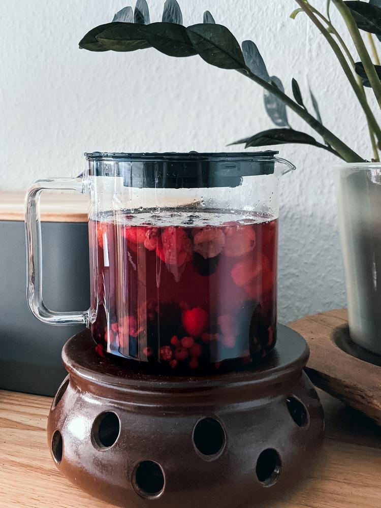 Eine Teekanne auf einem braunen Stövchen. In der Kanne sieht man verschiedene Beerenfrüchte in rotem Tee schwimmen.